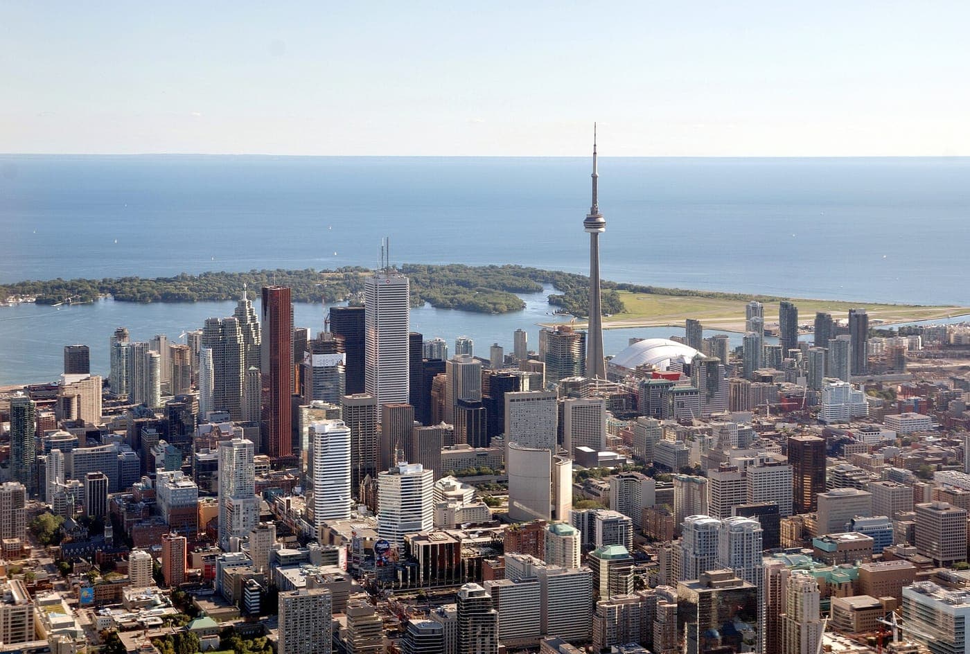 Aerial view of downtown Toronto's high-rise skyline with the CN Tower, the white Rogers Centre dome, the Toronto Islands and Lake Ontario under a clear sky