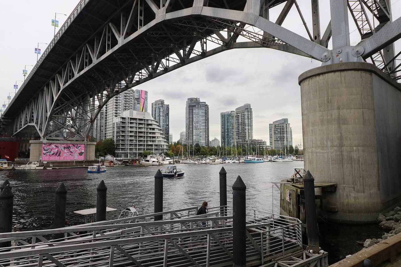 Underside of the Granville Street Bridge over False Creek, with a ferry dock, small boats, a marina and high-rise condo towers under an overcast sky