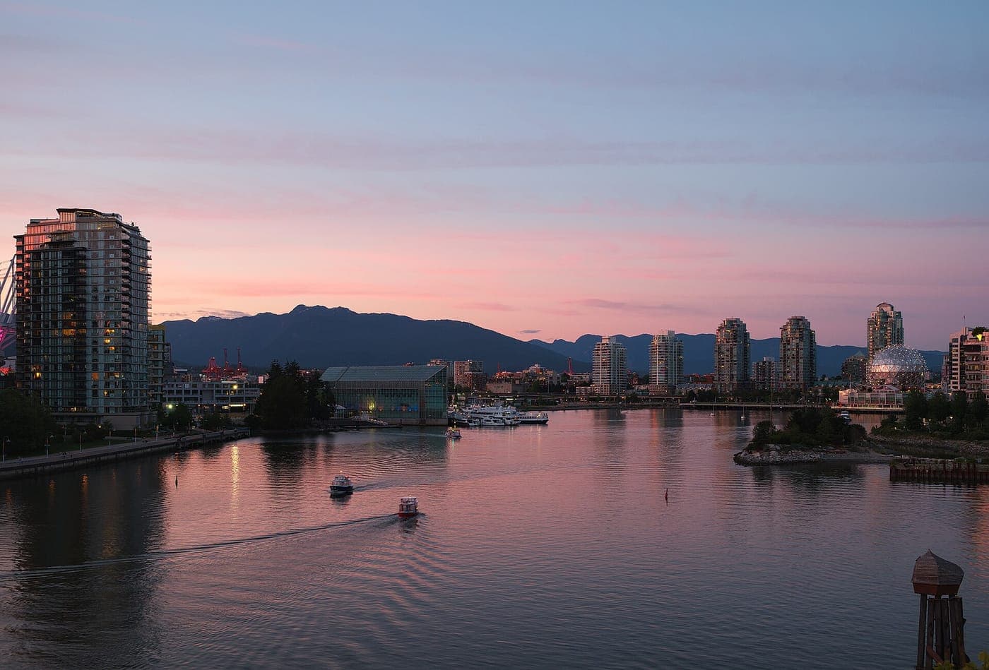 Pink dusk sky over False Creek as small ferries cross calm water, with high-rise condo towers, the Science World geodesic dome and dark North Shore mountains beyond