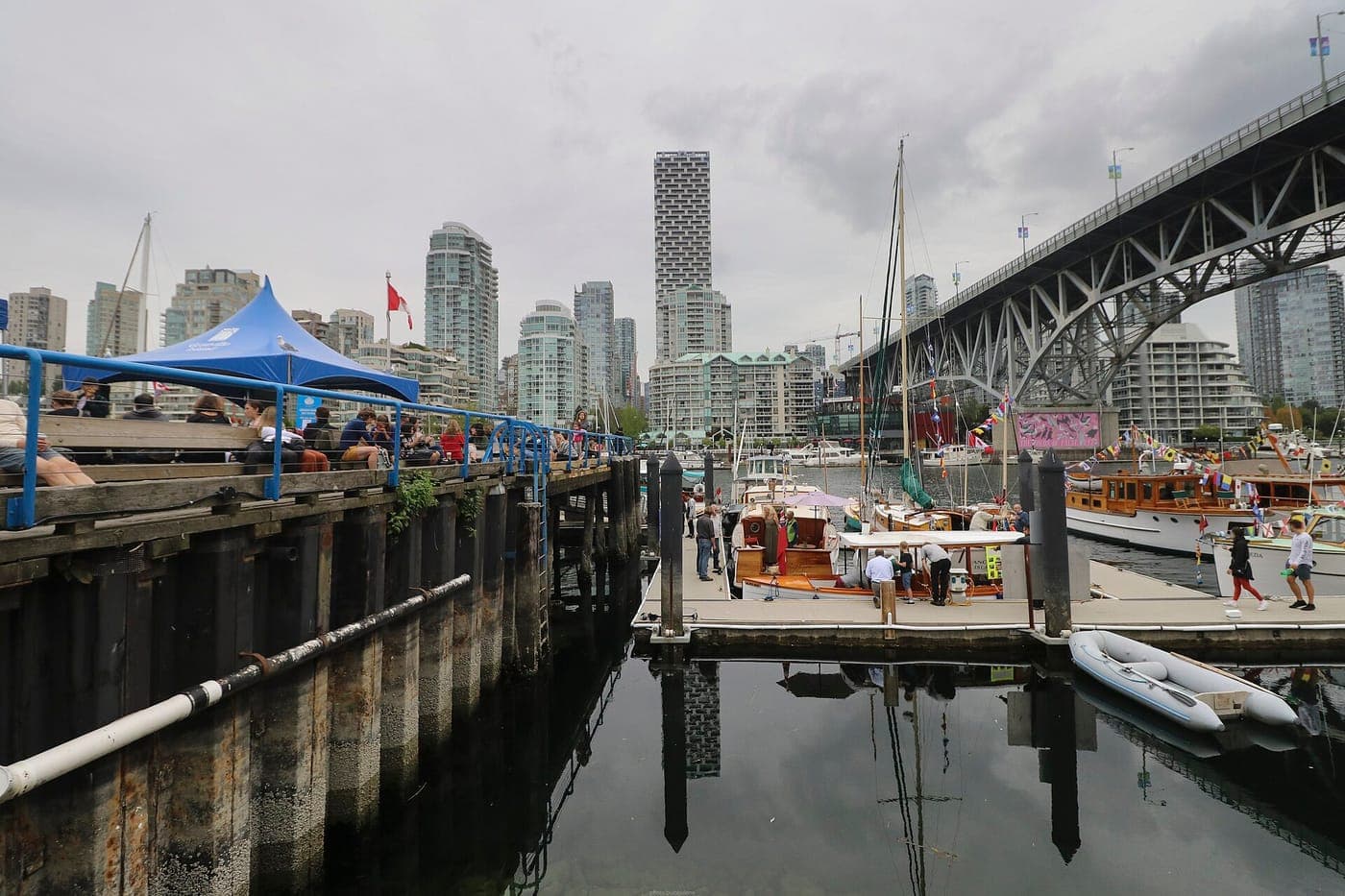 Wooden boats moored at a False Creek dock beneath the Granville Street Bridge, with people seated on a pier and downtown Vancouver condo towers under an overcast sky.