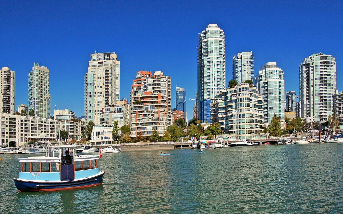 A small blue passenger ferry crosses False Creek in Vancouver, with glassy condo towers, mid-rise apartments and a marina lining the waterfront under a clear blue sky.