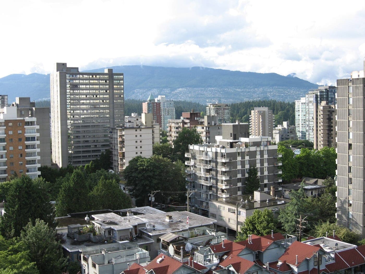Dense mix of mid-rise and high-rise apartment towers among leafy trees, with red-roofed townhouses in front and forested mountains under cloud behind