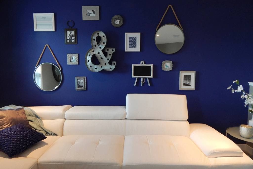 A white sectional sofa set against a deep blue wall arranged with framed pictures, two round rope-hung mirrors and a metal ampersand sign.