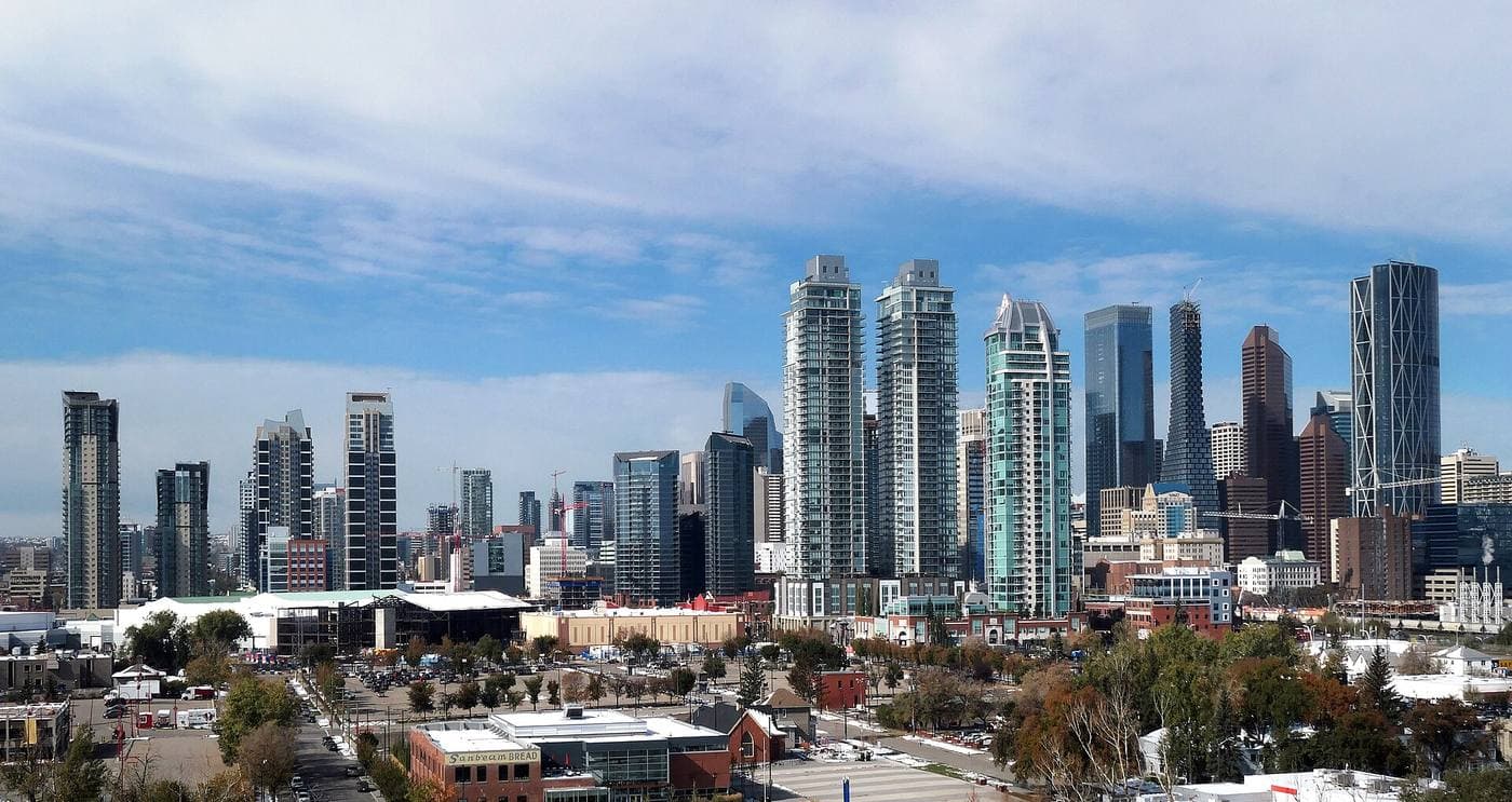 Clusters of glass residential towers and construction cranes rise above low-rise brick blocks and autumn trees at the edge of downtown Calgary.