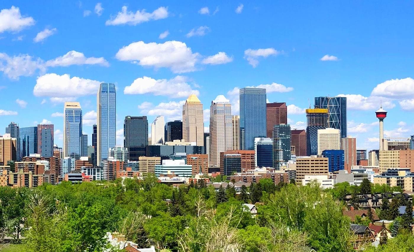 Downtown Calgary's high-rise cluster, including the red-topped Calgary Tower, seen above mature green trees and low-rise homes under a bright blue sky.