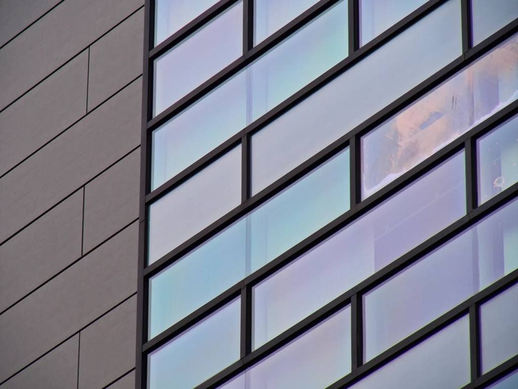 Angled view of a modern tower's glass curtain wall, with pale blue panes in dark frames beside grey cladding.
