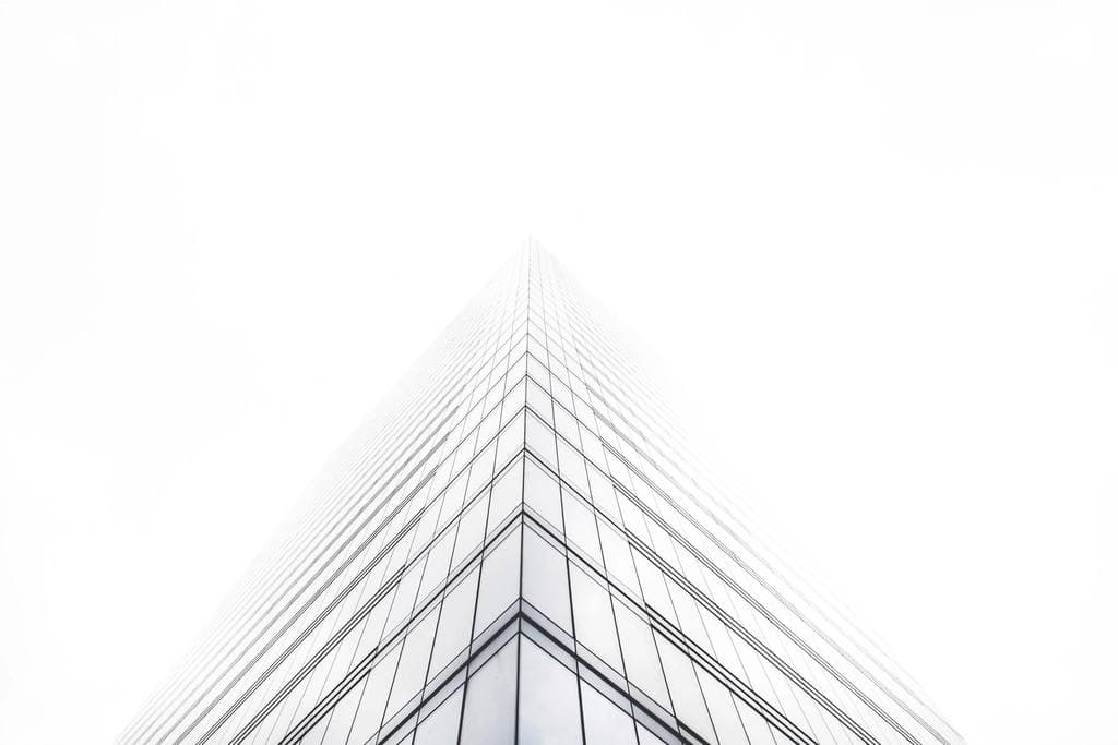 Looking straight up the mirrored corner of a modern glass office tower as it recedes into a bright, featureless white sky.