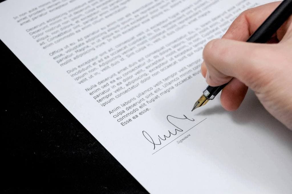 A hand holds a fountain pen and signs the signature line at the foot of a printed agreement resting on a dark desktop.