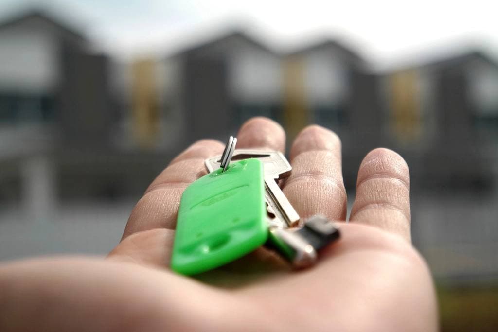 A set of house keys with a bright green plastic fob resting in an open palm, with softly blurred townhouses behind.