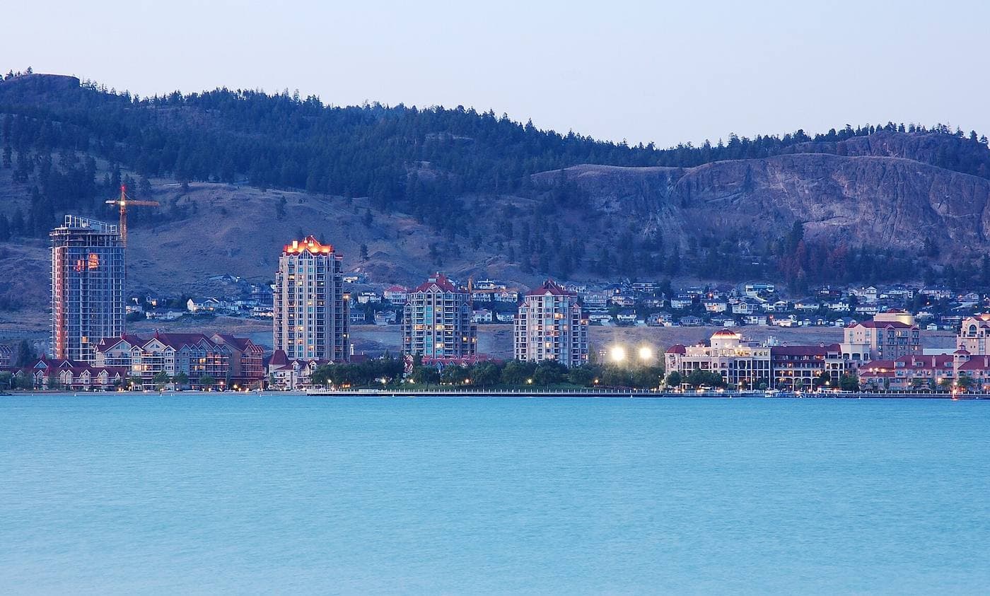 Downtown highrise towers and lakefront buildings lit at dusk across Okanagan Lake, a construction crane above them and dry pine hillsides behind.