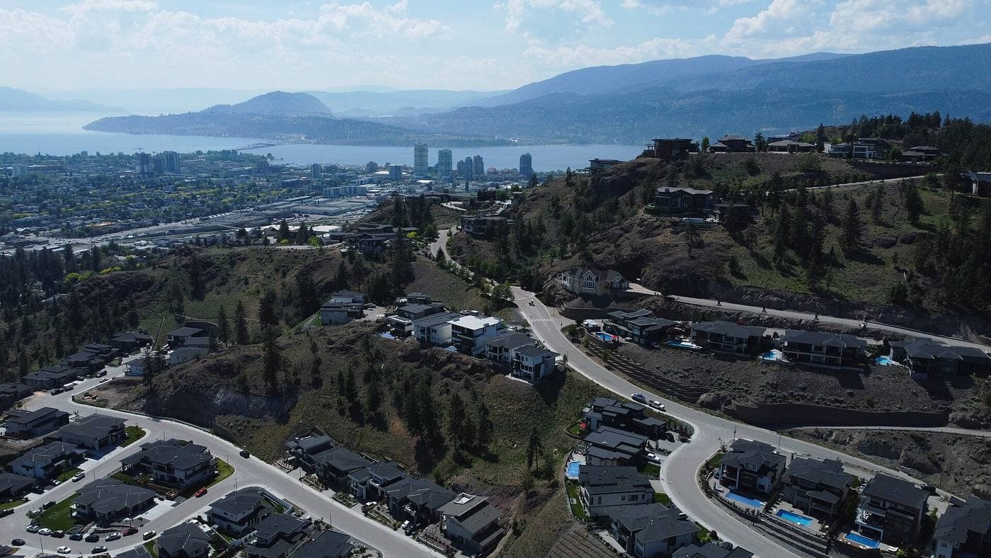 Aerial view over hillside subdivisions of modern homes and curving streets, with a downtown highrise cluster and Okanagan Lake beyond.