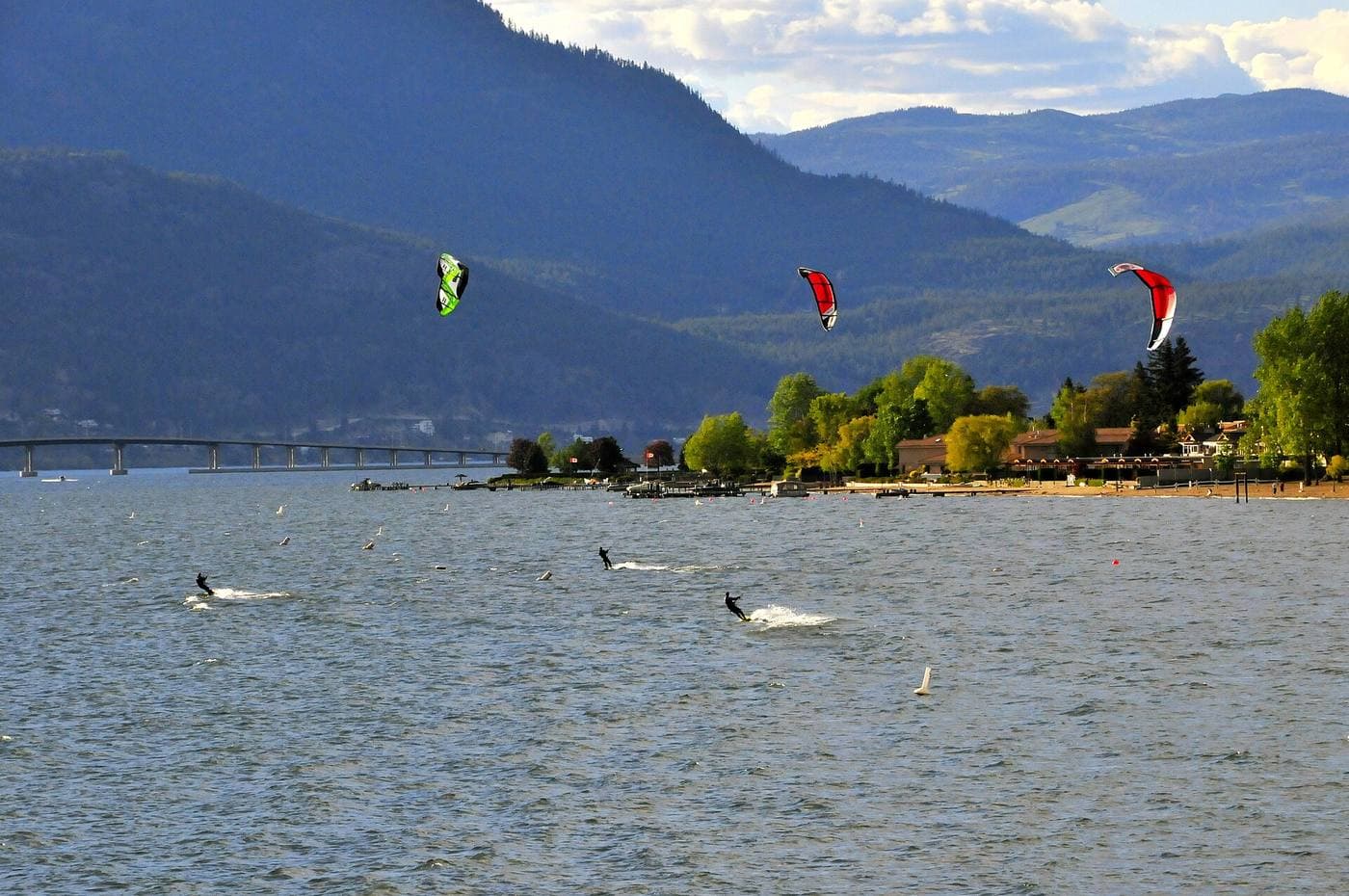 Kiteboarders skim Okanagan Lake in front of a long low floating bridge, a tree-lined beach with lakeside homes and forested hills beyond.