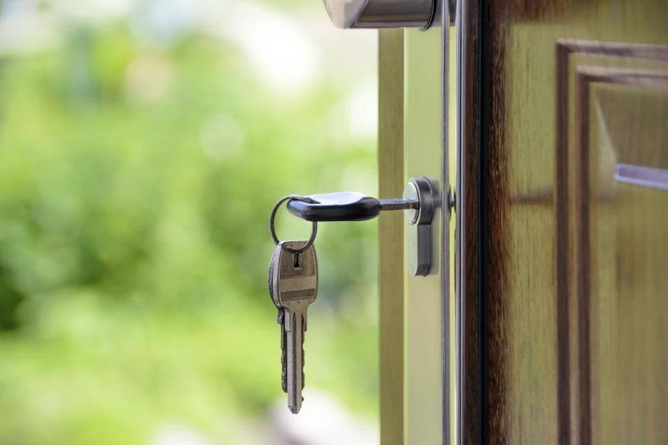 A set of keys left hanging in the lock of an open front door, with green foliage blurred in the background.