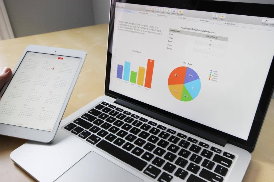 An open laptop showing colourful bar and pie charts sits on a wooden desk while a hand holds a tablet displaying a full-year calendar.