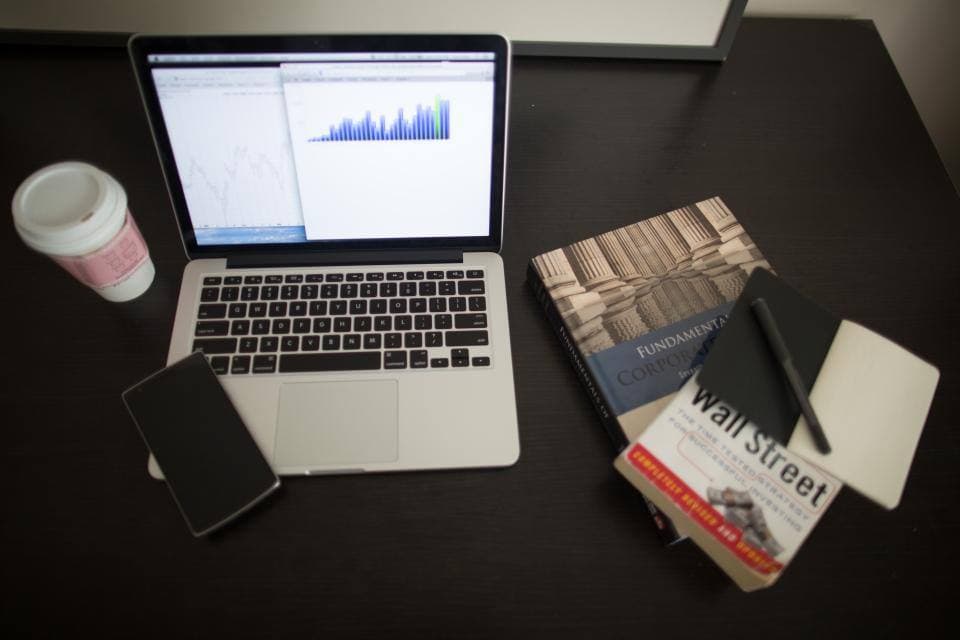 An open laptop showing a spreadsheet and a blue bar chart sits on a dark desk beside a takeaway coffee cup, a phone, a notepad and two finance textbooks.