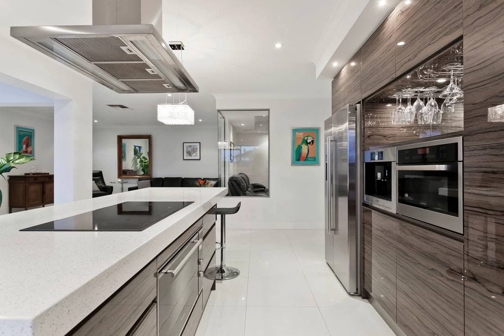 A modern kitchen with a white quartz island and cooktop, wood-panelled cabinetry, stainless steel appliances and stemware hanging above the counter.