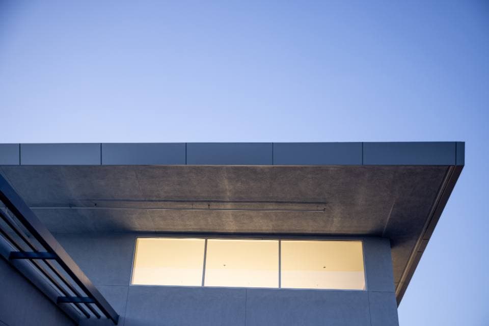 Deep flat roof overhang of a modern concrete building seen from below, with a band of warmly lit windows glowing against a deep blue evening sky.
