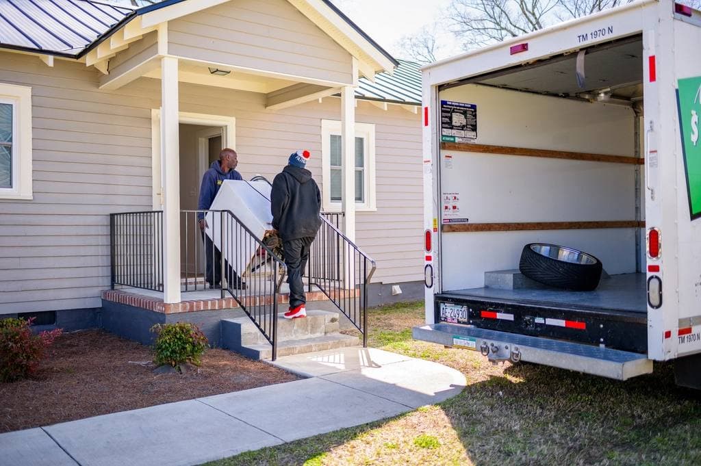 Two movers carry a large white appliance up the front steps of a clapboard house while the open rear of a moving truck waits on the lawn.