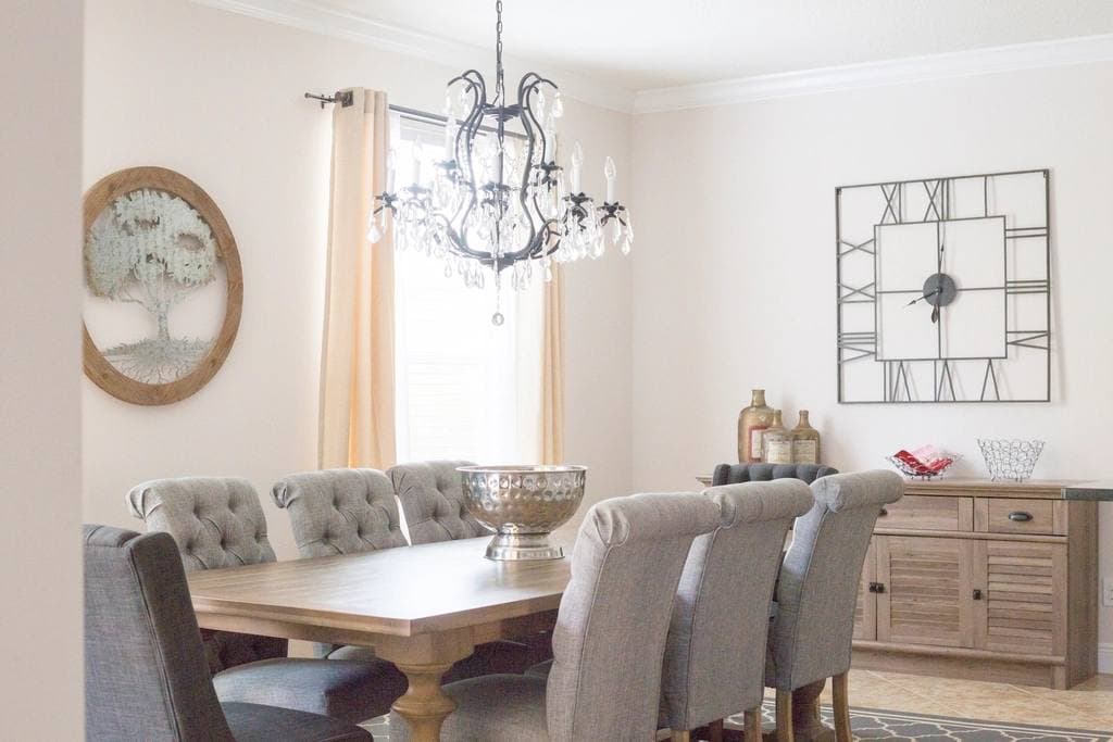 A pale wood dining table ringed by tufted grey chairs under a black crystal chandelier, with a sideboard and large wall clock behind.