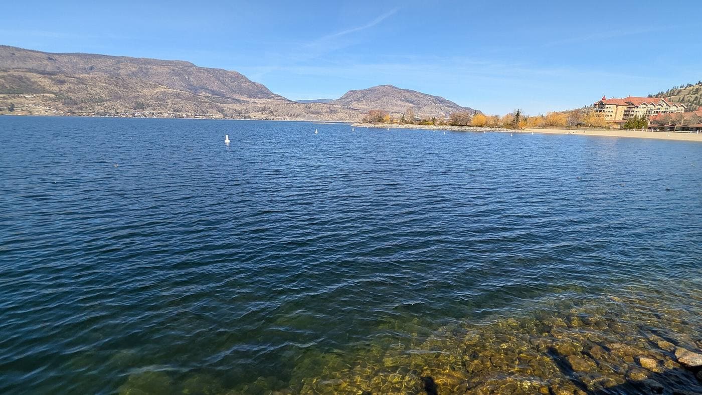 Clear blue Okanagan Lake water over a pebbled shallow edge, with a sandy beach, red-roofed lakefront condo buildings and dry brown hills behind.