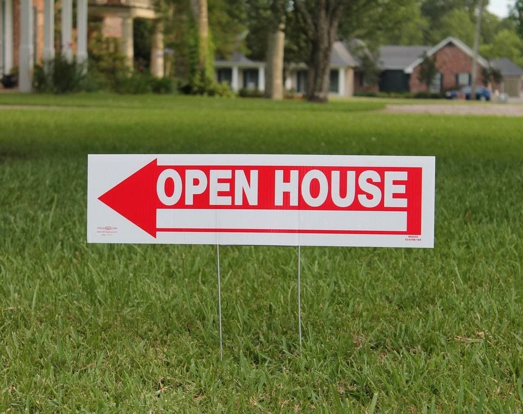 A red and white arrow sign reading Open House staked into a freshly mown front lawn, with suburban houses blurred behind.