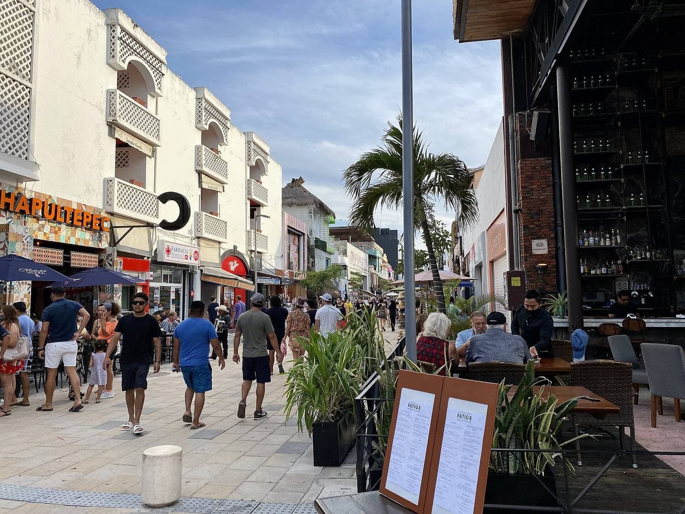 Pedestrians walk a palm-lined pedestrian shopping street past whitewashed balconied buildings, a pharmacy and open-air bar patios under a blue sky