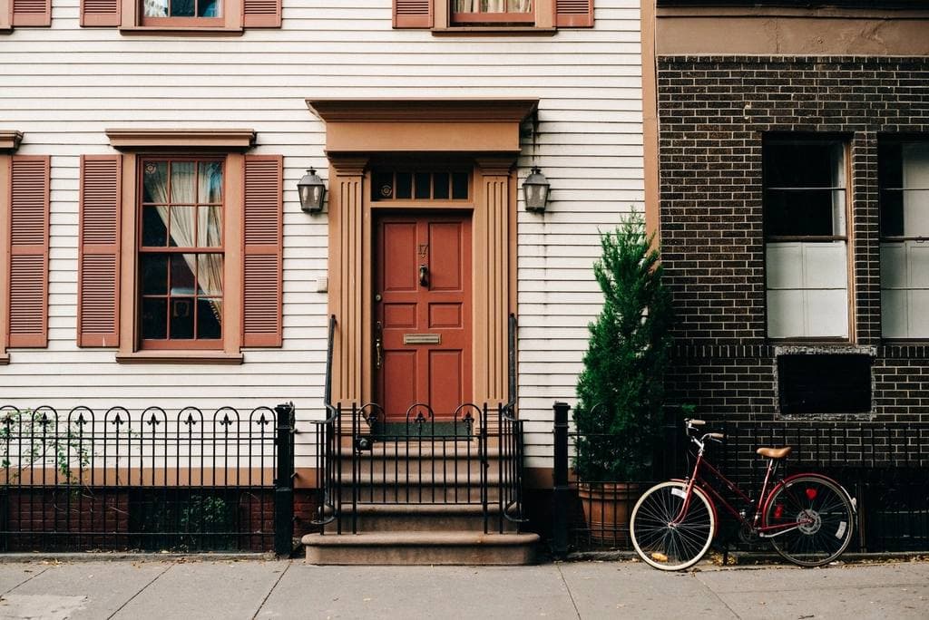 A red panelled front door with carriage lamps on a white clapboard rowhouse, with a bicycle leaning against the brick building next door.