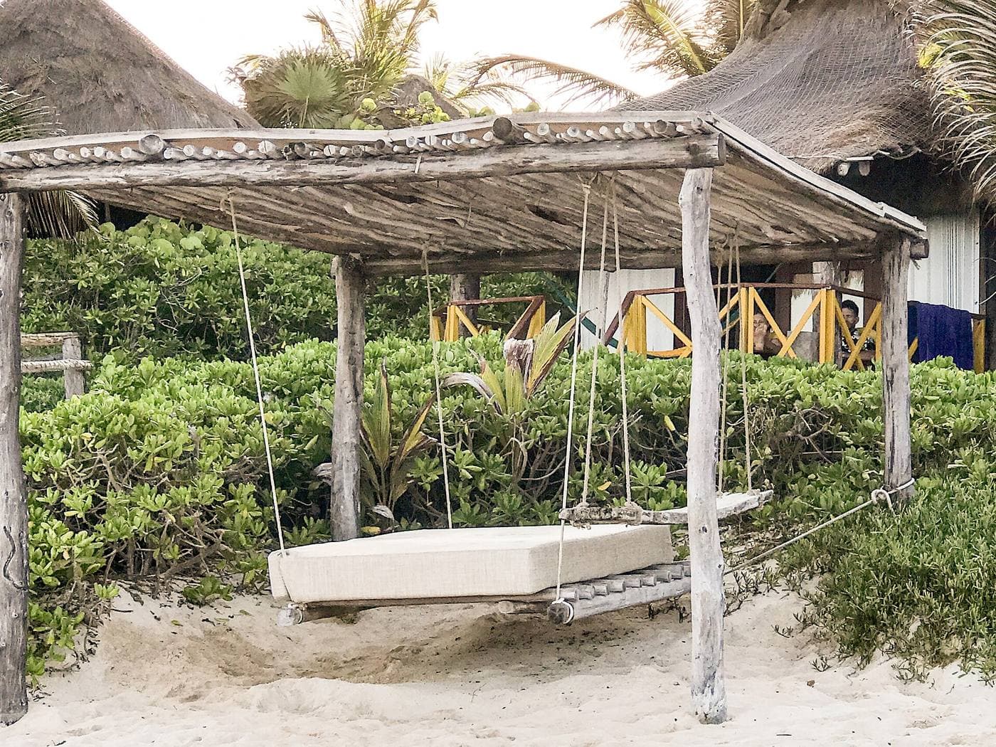 A shaded beach cabana with a hanging daybed among sea-grape trees at a Riviera Maya resort.