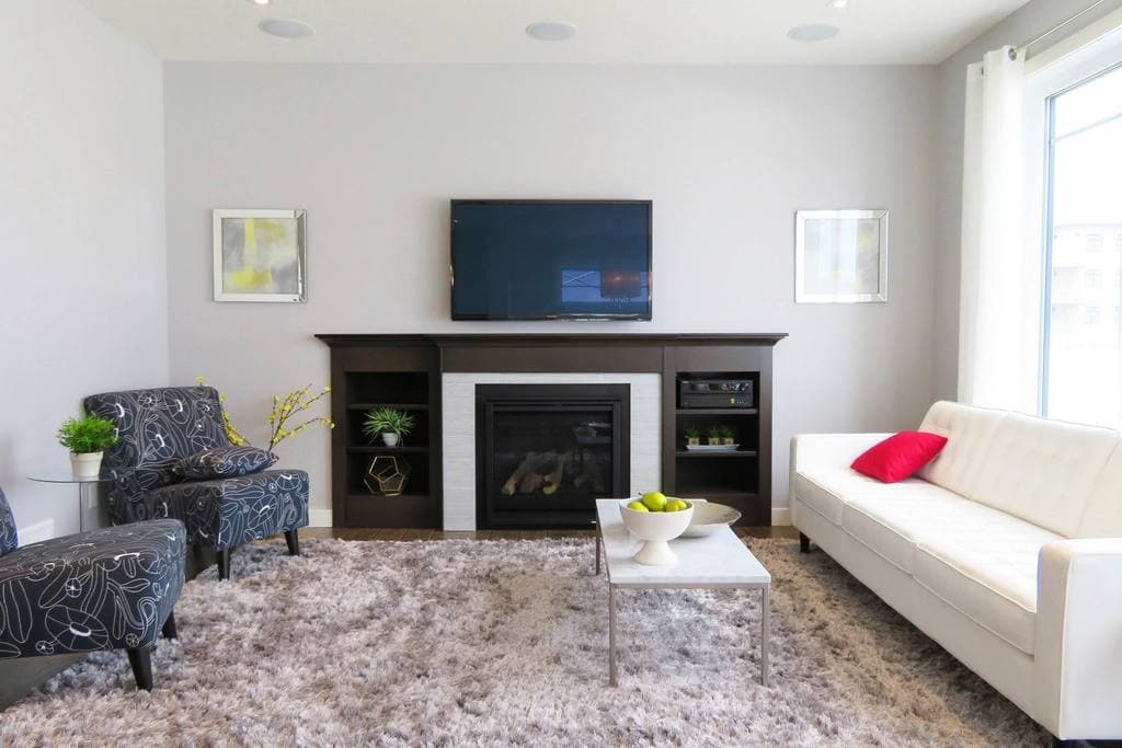 Staged living room with a white leather sofa, patterned armchairs, a fireplace below a mounted television and a deep shag rug.