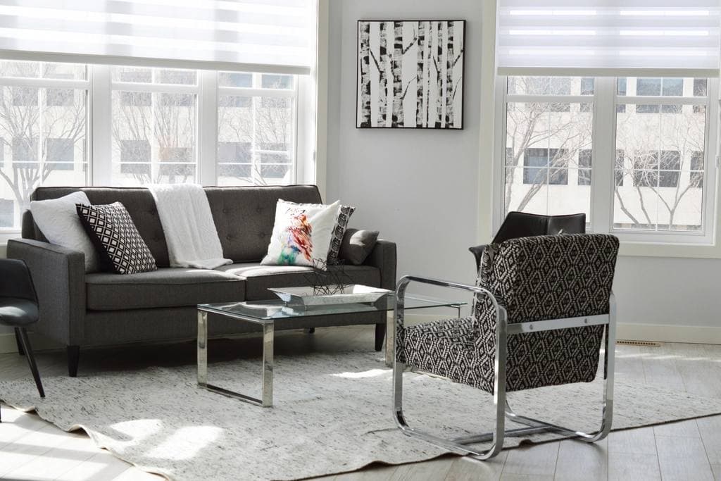 Sunlit living room with a grey tufted sofa, patterned cushions, a glass coffee table and a woven armchair on a pale rug.