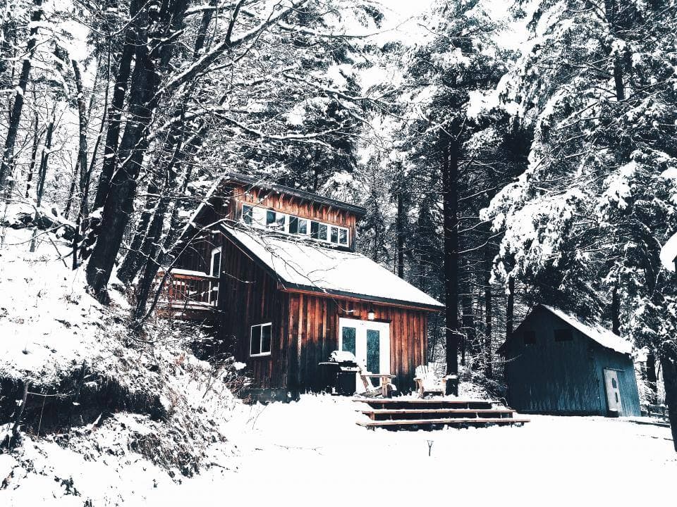 A dark timber cabin with a steep snow-covered roof sits among tall evergreens, its wooden steps and deck chairs buried in deep snow.