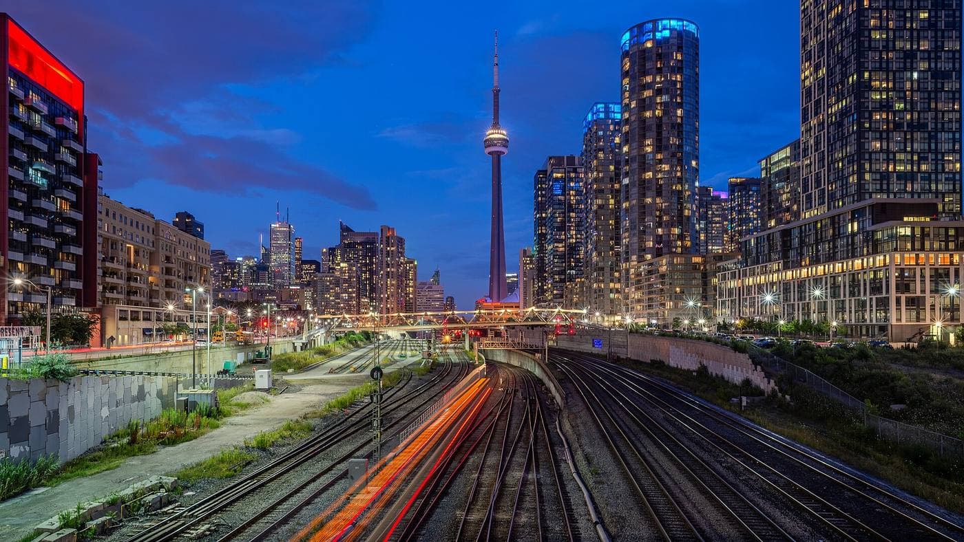 Rail lines curve toward downtown Toronto at blue hour, with the CN Tower and brightly lit condominium towers rising on both sides of the corridor.