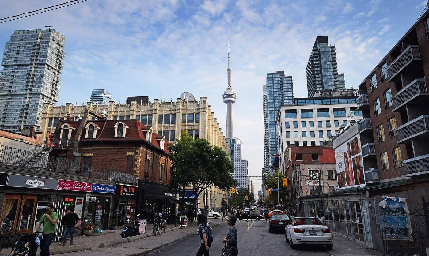 Low-rise shops and Victorian brick buildings line a downtown Toronto street, with glass condo towers and the CN Tower rising behind the intersection.