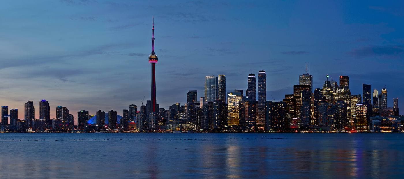 Toronto's downtown skyline lit at dusk across calm lake water, the CN Tower glowing pink among clusters of office and condo towers.