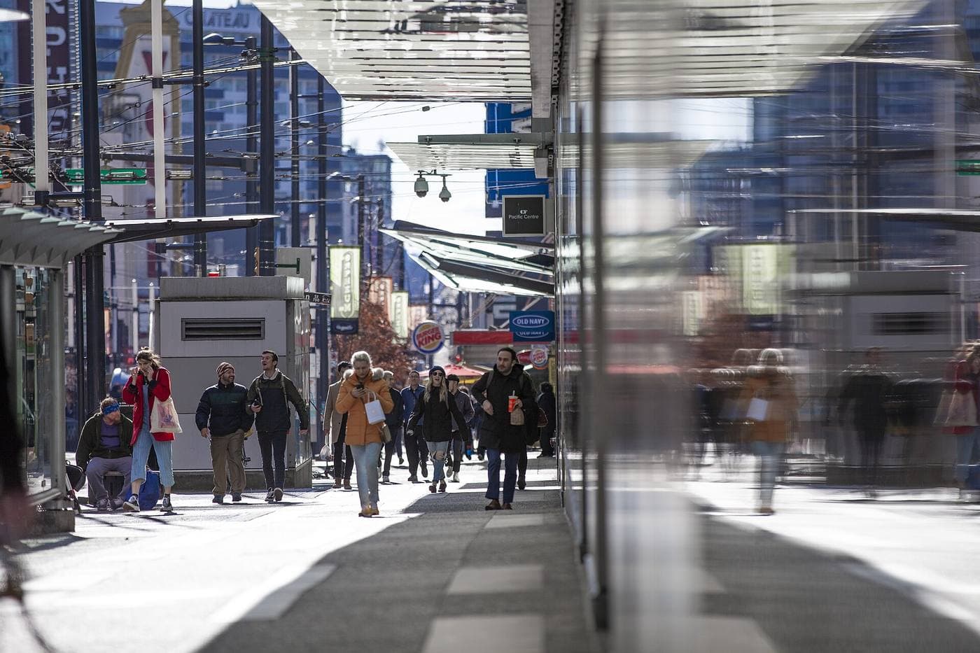 Pedestrians walk a sunlit downtown Vancouver sidewalk past CF Pacific Centre and Old Navy signs beneath glass canopies and trolley wires.