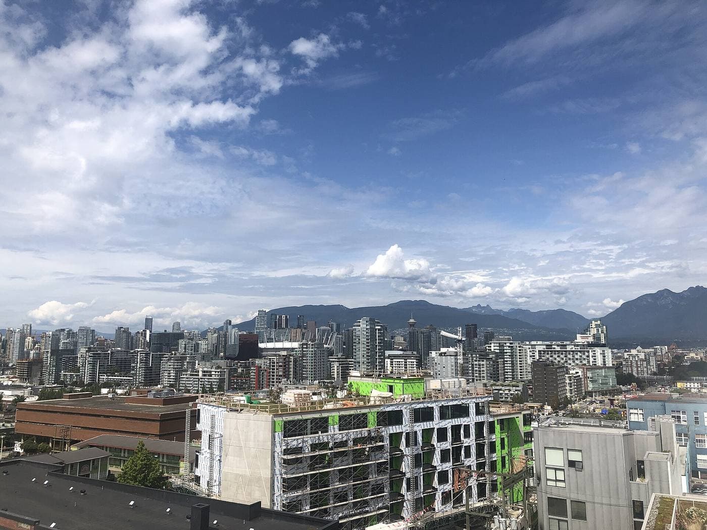 A mid-rise apartment block under construction in the foreground, with downtown Vancouver's towers, BC Place and the North Shore mountains beyond.