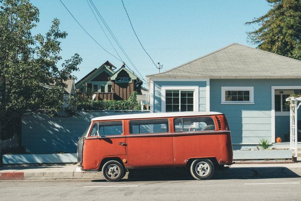 A vintage orange-and-white camper van parked at the curb in front of a pale blue clapboard bungalow under a clear sky.