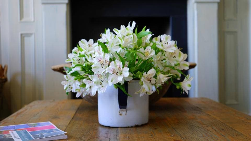 White alstroemeria blooms in a chipped enamel jug on a rustic plank table in front of a painted fireplace surround.