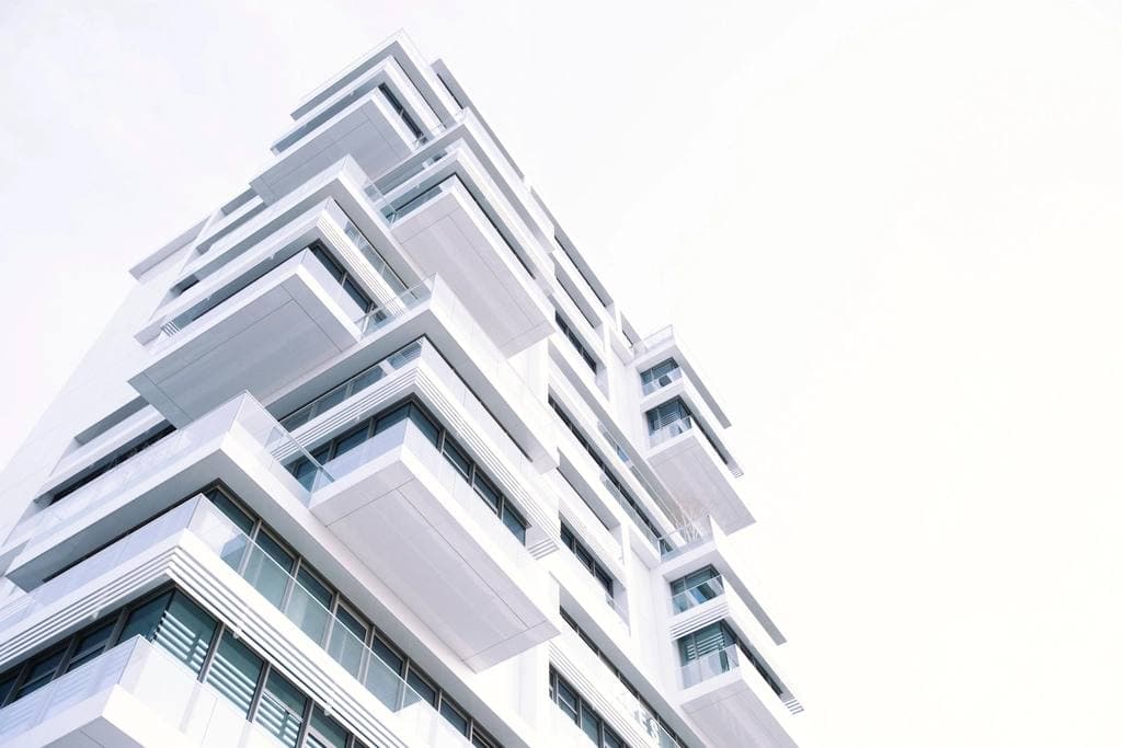 Low-angle view of a bright white contemporary apartment tower with stacked, glass-railed balconies against a pale sky.