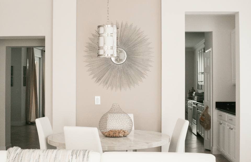 A small round white dining table with upholstered white chairs below a sunburst mirror and lantern pendant, beside an open kitchen doorway.
