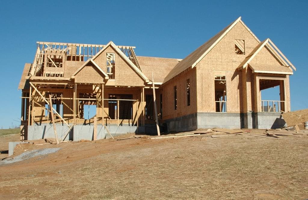 A half-built two-storey house framed in bare lumber and plywood sheathing stands on a dirt lot under a clear blue sky.