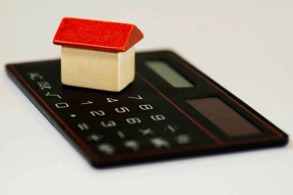 A small wooden toy house with a bright red roof resting on a slim black solar calculator against a plain white surface.