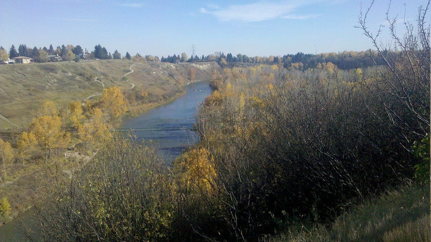 A river winds through a valley of autumn-yellow trees beneath a grassy bluff with walking paths, with houses and evergreens along the top of the ridge under a blue sky.