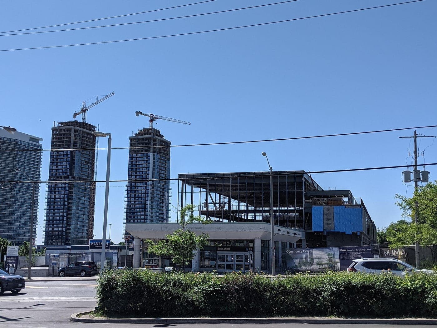 Two high-rise condo towers under construction with cranes stand behind a steel-framed building going up next to a subway station entrance, with a hedge and street in front.