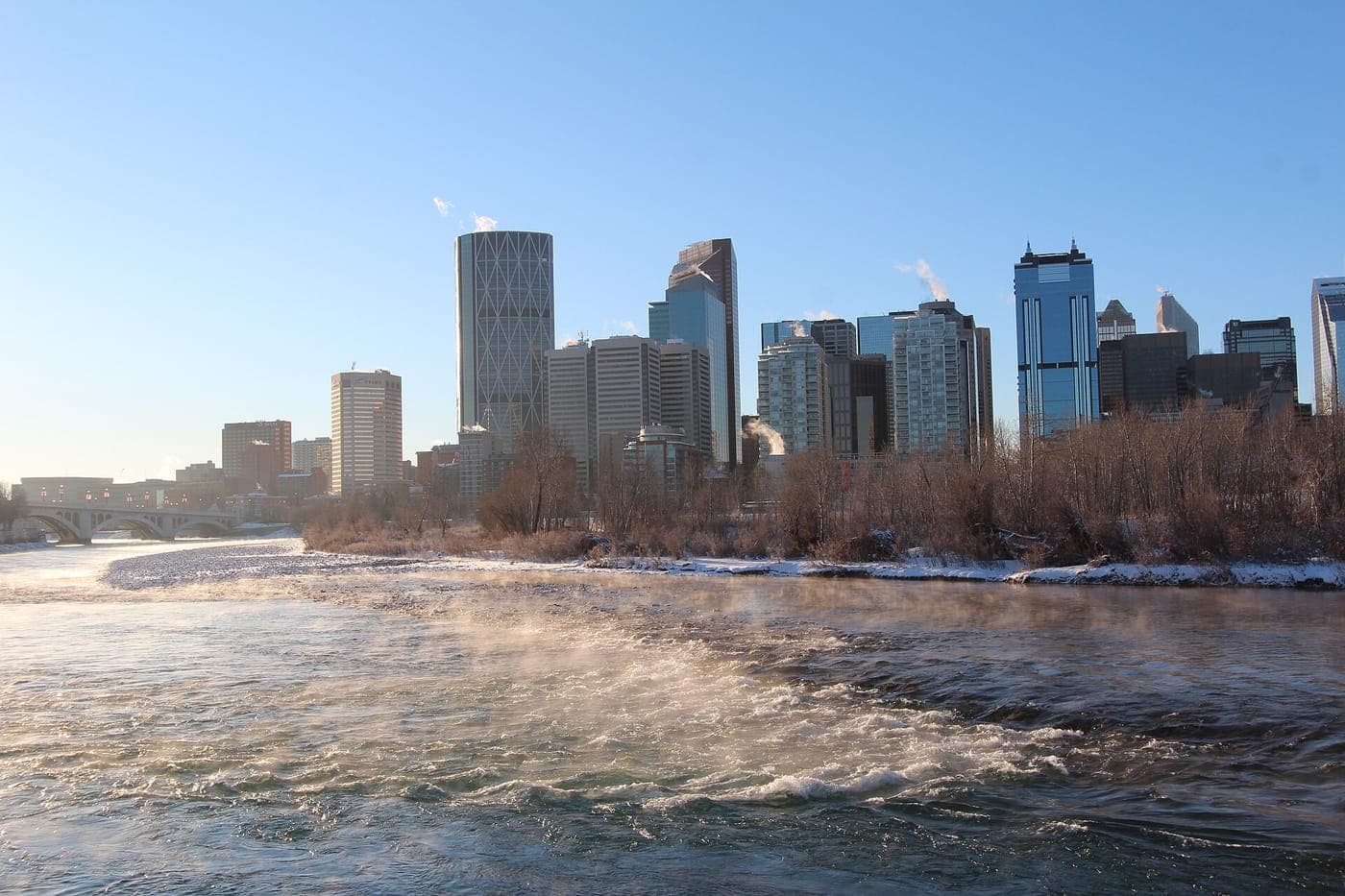Downtown Calgary skyline, including The Bow tower, rises behind leafless trees and snowy banks as mist drifts off the Bow River on a clear winter morning.