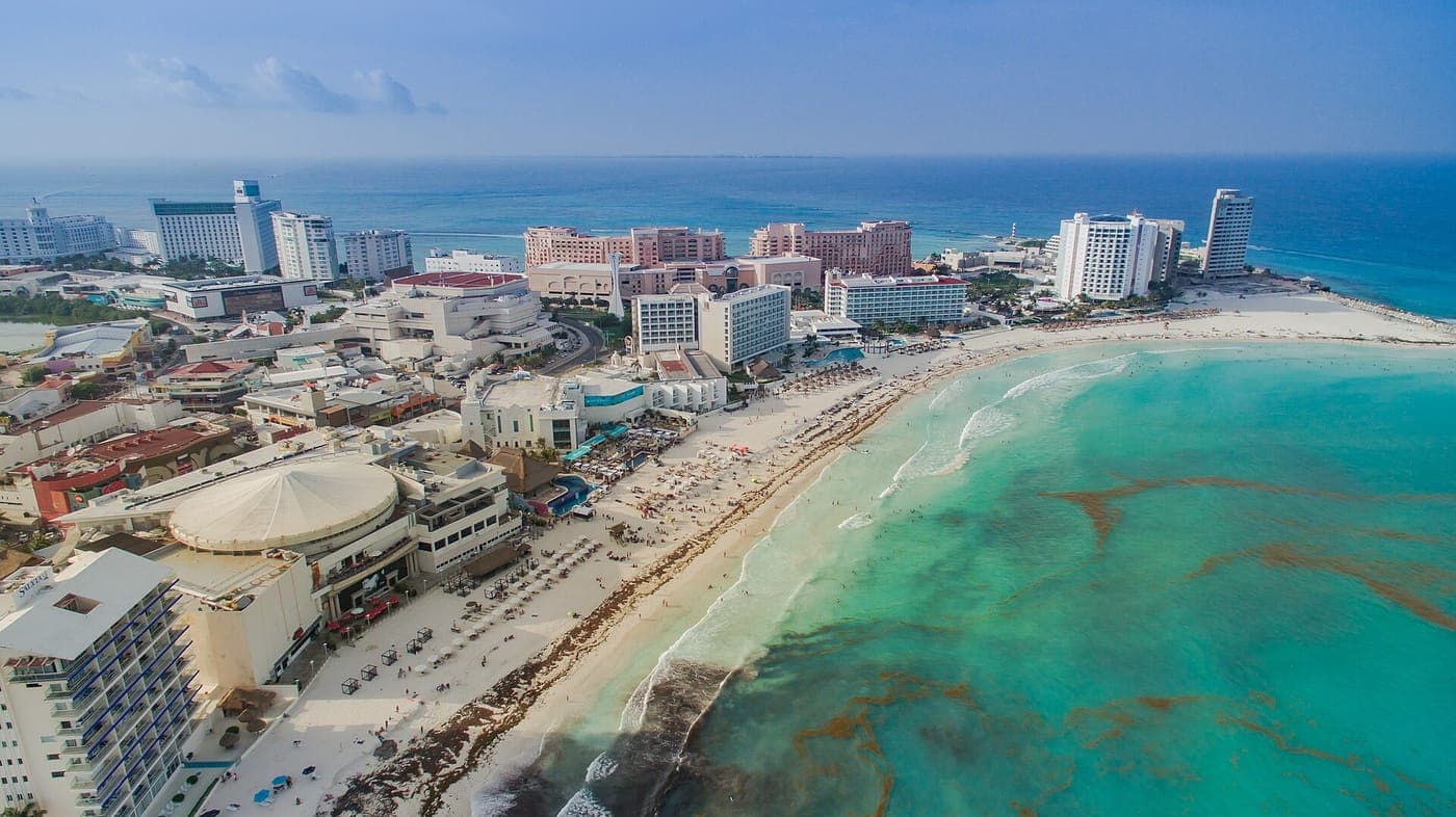 Aerial view of a curving white-sand beach lined with resort hotels and high-rises, turquoise Caribbean water with patches of seaweed, and open sea beyond.