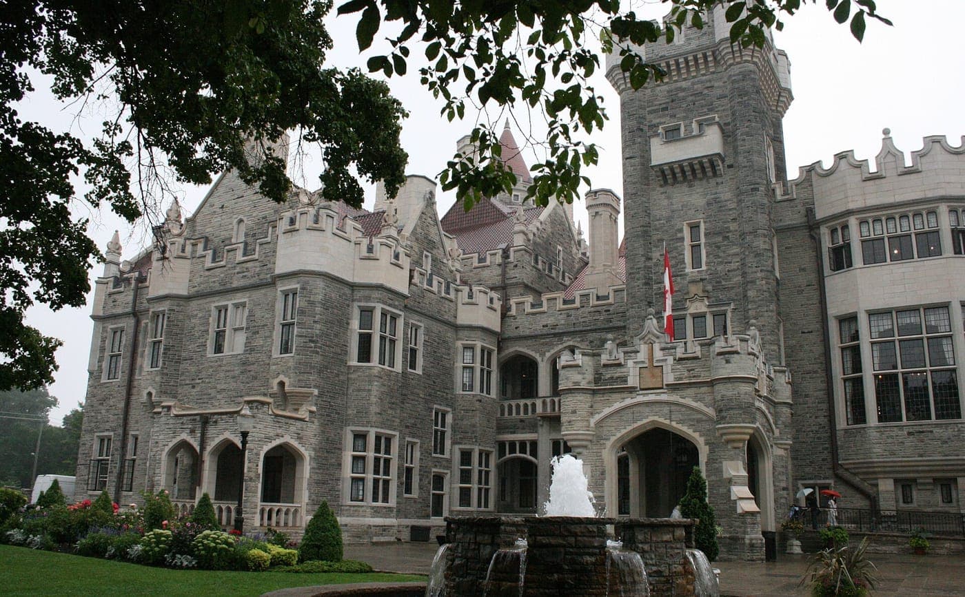 Casa Loma's grey stone Gothic Revival castle with turrets and battlements, a Canadian flag, and a stone fountain and flower beds in front on an overcast day.