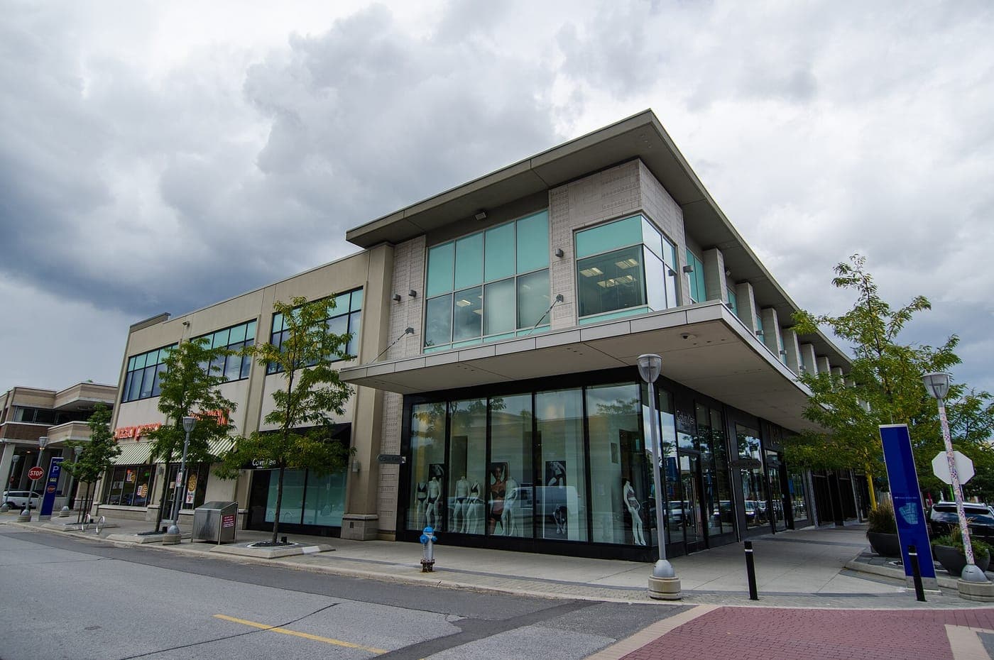 Modern two-storey retail building with large glass storefront windows displaying mannequins, young street trees, a wide sidewalk and a brick crosswalk under a cloudy sky