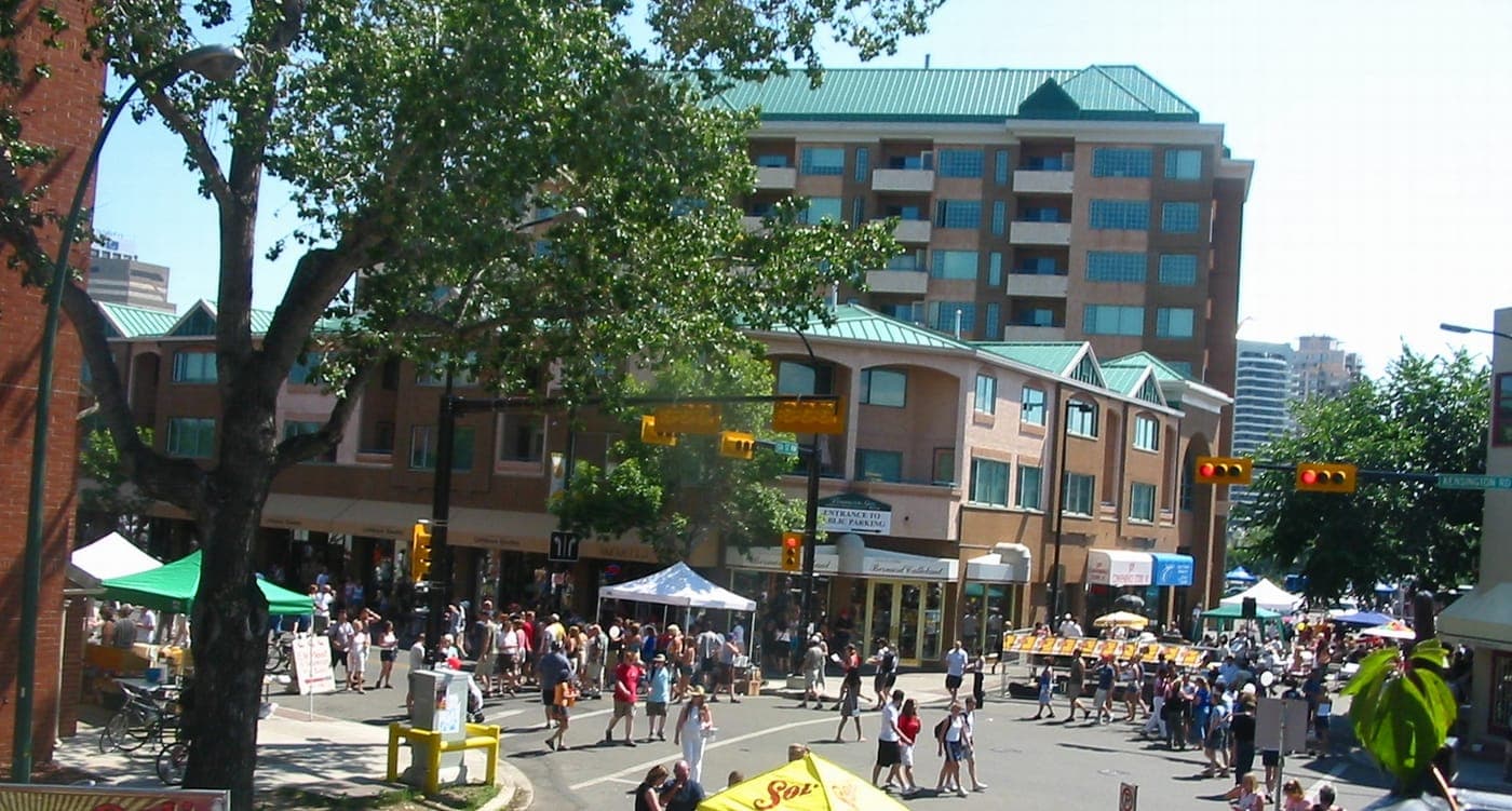 Crowds stroll a closed-off street during a summer festival, with vendor tents, traffic lights, leafy trees and a green-roofed brick mixed-use condo building behind
