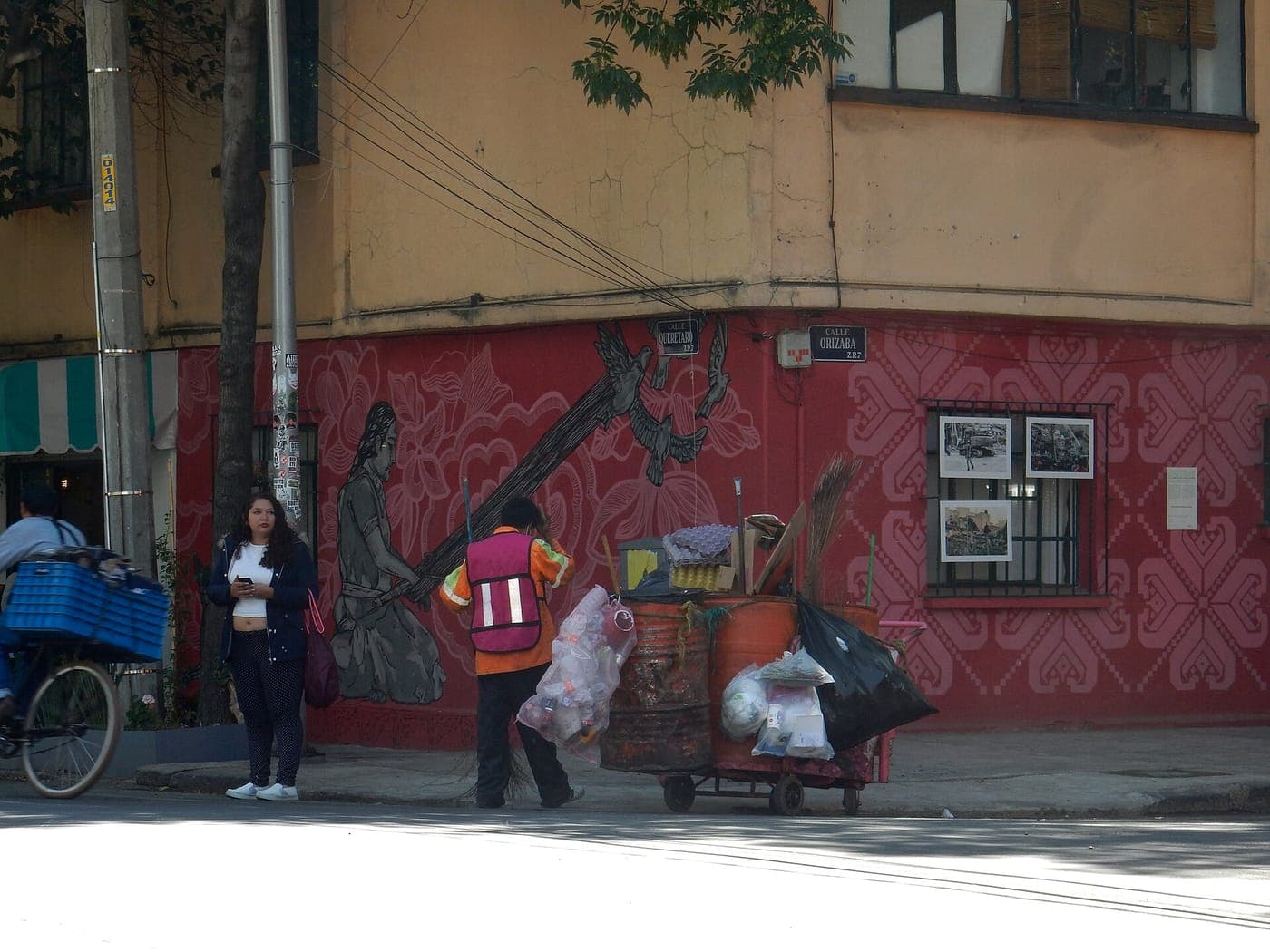 Street corner with a red patterned mural wall below a yellow building, where a street sweeper in a safety vest stands by a cart of barrels and bags while a woman waits on the sidewalk